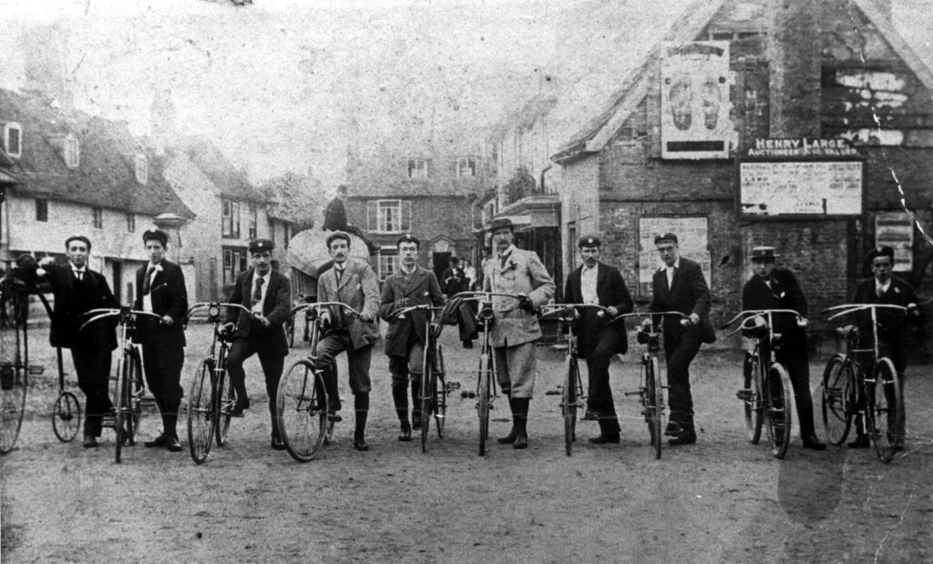 A row of men in old fashion clothing standing next to old-fashioned cycles including one penny farthing, black and white photo
