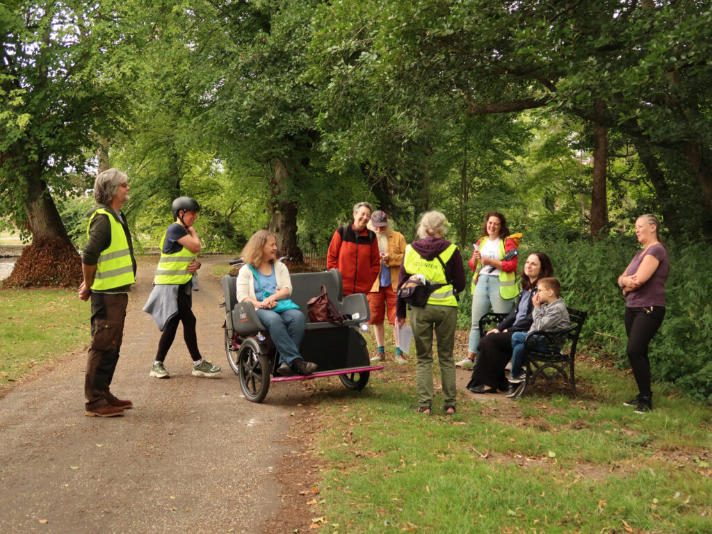 walkers, one on rickshaw, listening to speaker by river under lime trees