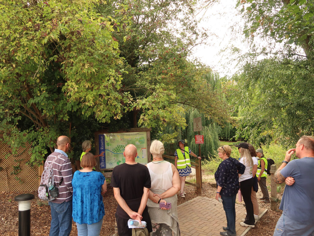 walkers listening to speaker by nature reserve panel, river and trees behind